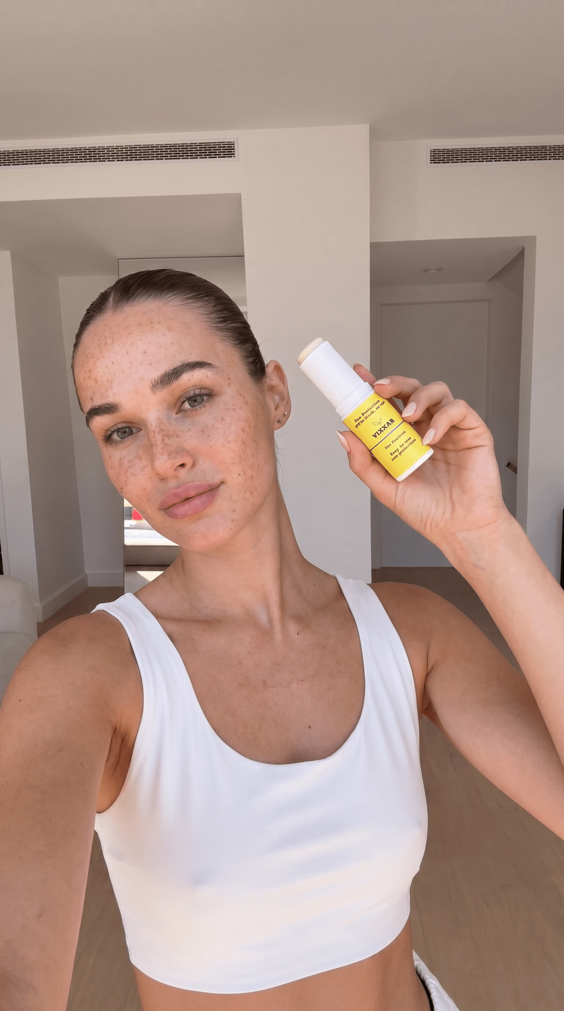 A woman in a white sports bra holds up a small yellow spf stick with a white cap, showing the front label of the product in a bright indoor setting.