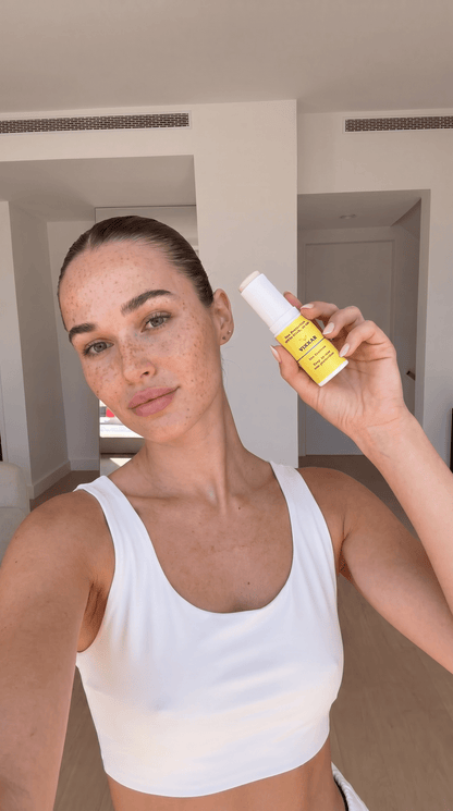 A woman in a white sports bra holds up a small yellow spf stick with a white cap, showing the front label of the product in a bright indoor setting.
